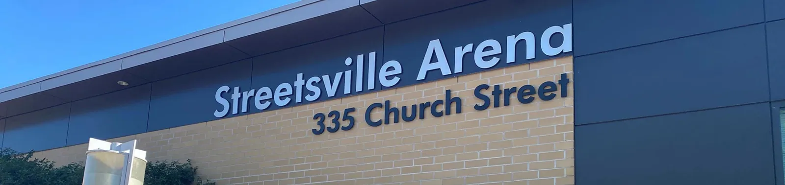 Angled view of the building's exterior wall, with a large sign reading "Streetsville Arena / 335 Church Street" mounted on the dark cladding above tan brick. — Vic Johnston Community Centre