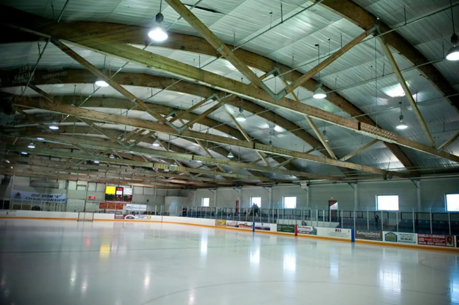 Empty ice sheet from ice level, showing the full rink boards, glass, a scoreboard, sponsor boards, and an arched wood-truss roof overhead. — Vic Johnston Community Centre