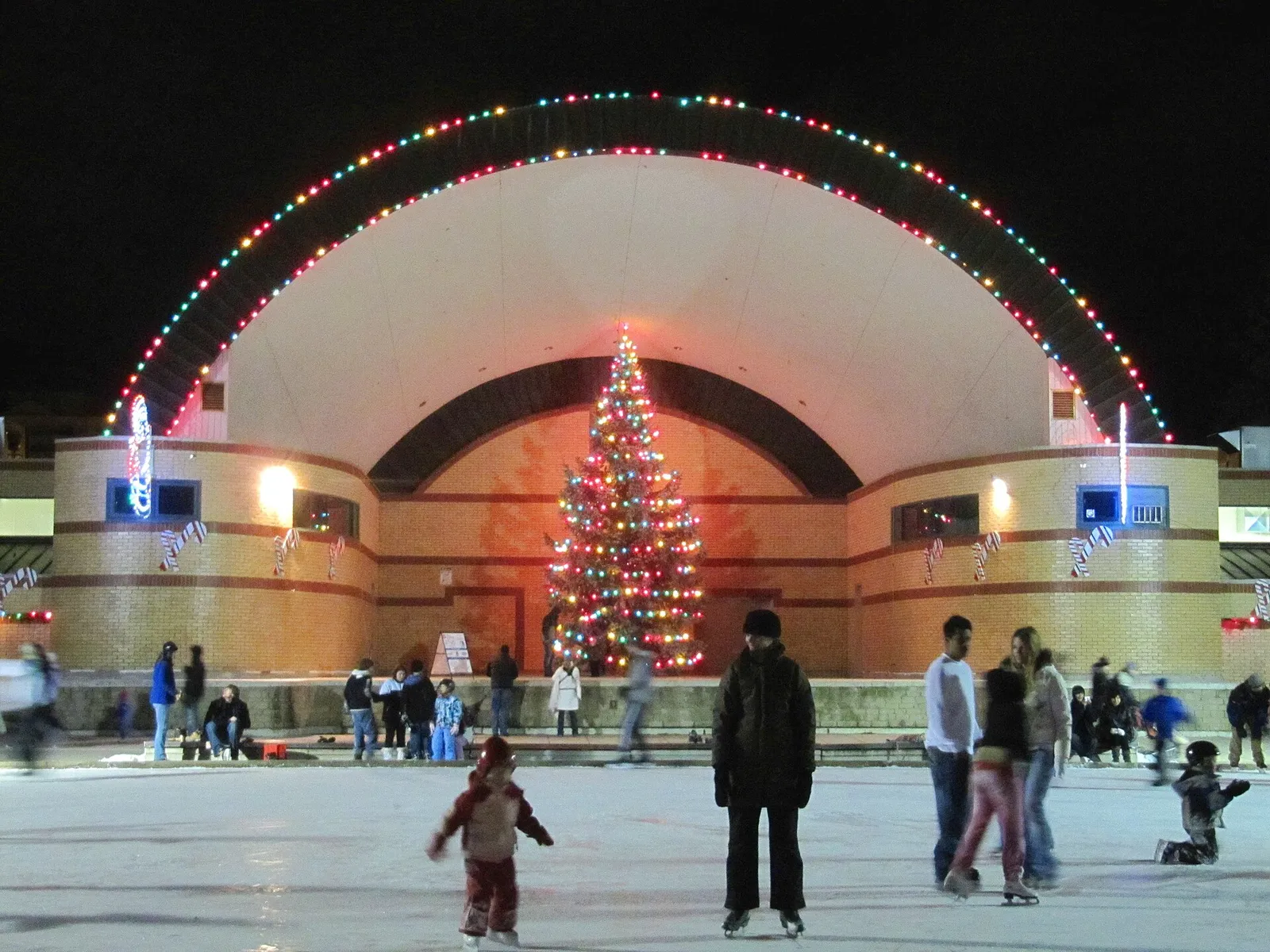 La pista de Victoria Park en London (Ontario), con patinadores frente al kiosco de música iluminado y un alto árbol de Navidad. — Victoria Park Outdoor Rink (London)