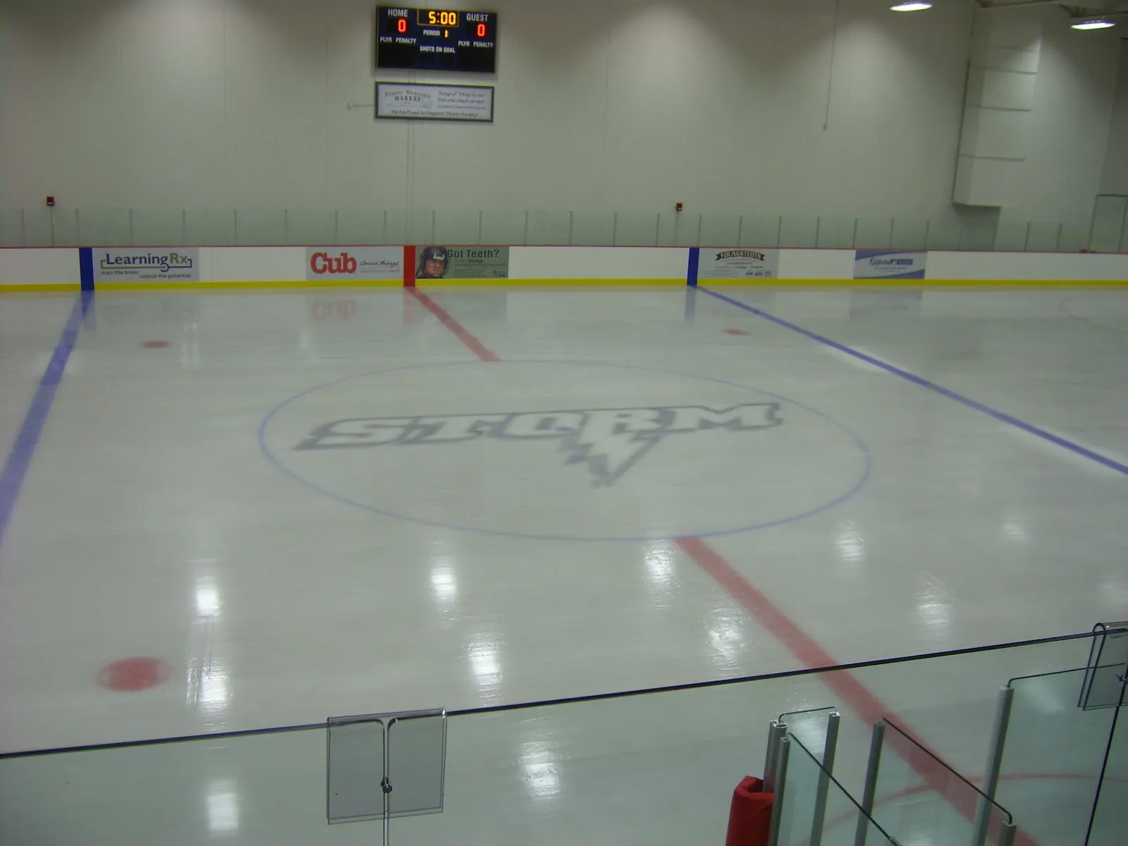 An empty sheet seen from behind the glass at one end, the Chanhassen Storm logo at centre ice under the scoreboard. — Victoria Recreation Center Ice Arena