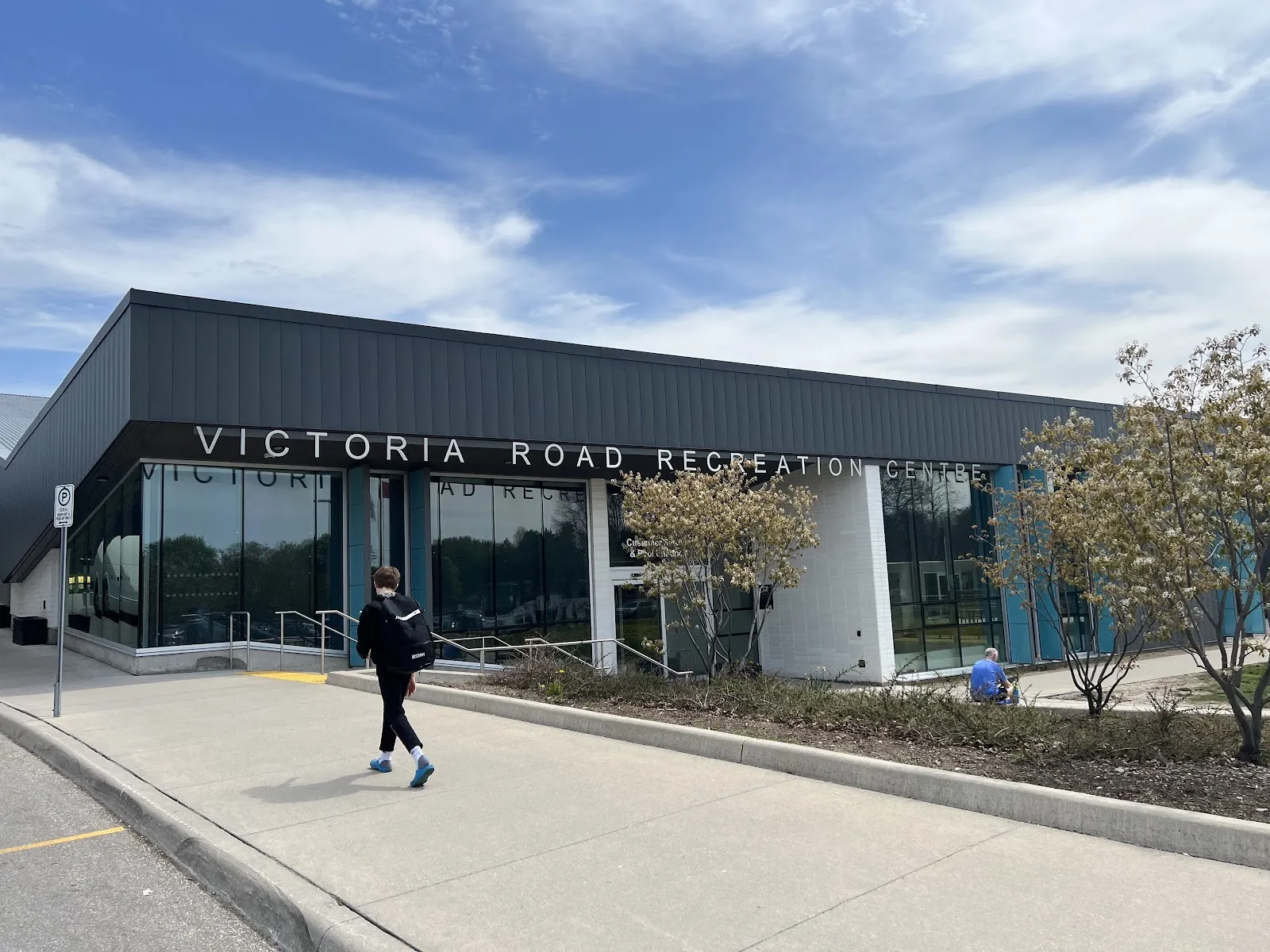 El Victoria Road Recreation Centre, con su nombre en letras plateadas a lo largo de una franja metálica oscura sobre altos muros de vidrio, y árboles junto al andador. — Victoria Road Recreation Centre