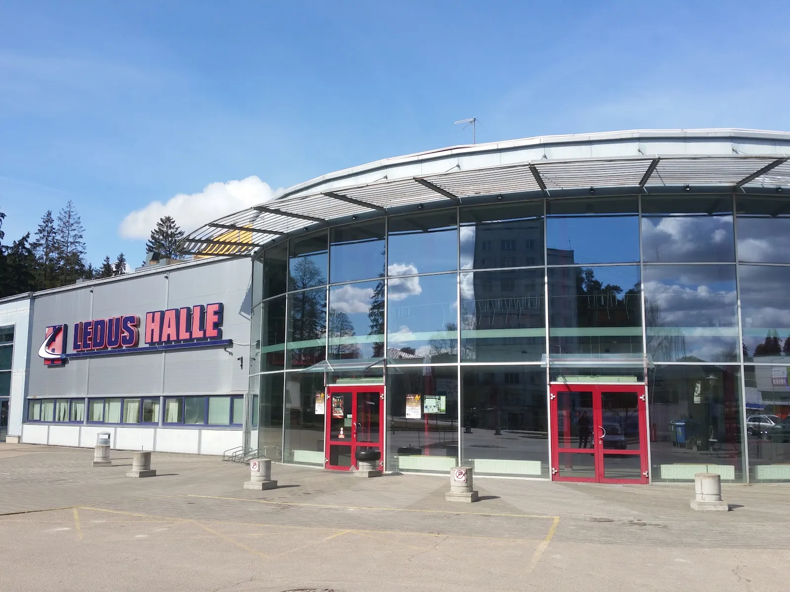 The front of the Vidzemes ice hall, a grey building with a curved glazed entrance hall and the red LEDUS HALLE lettering. — Vidzemes Ledus halle