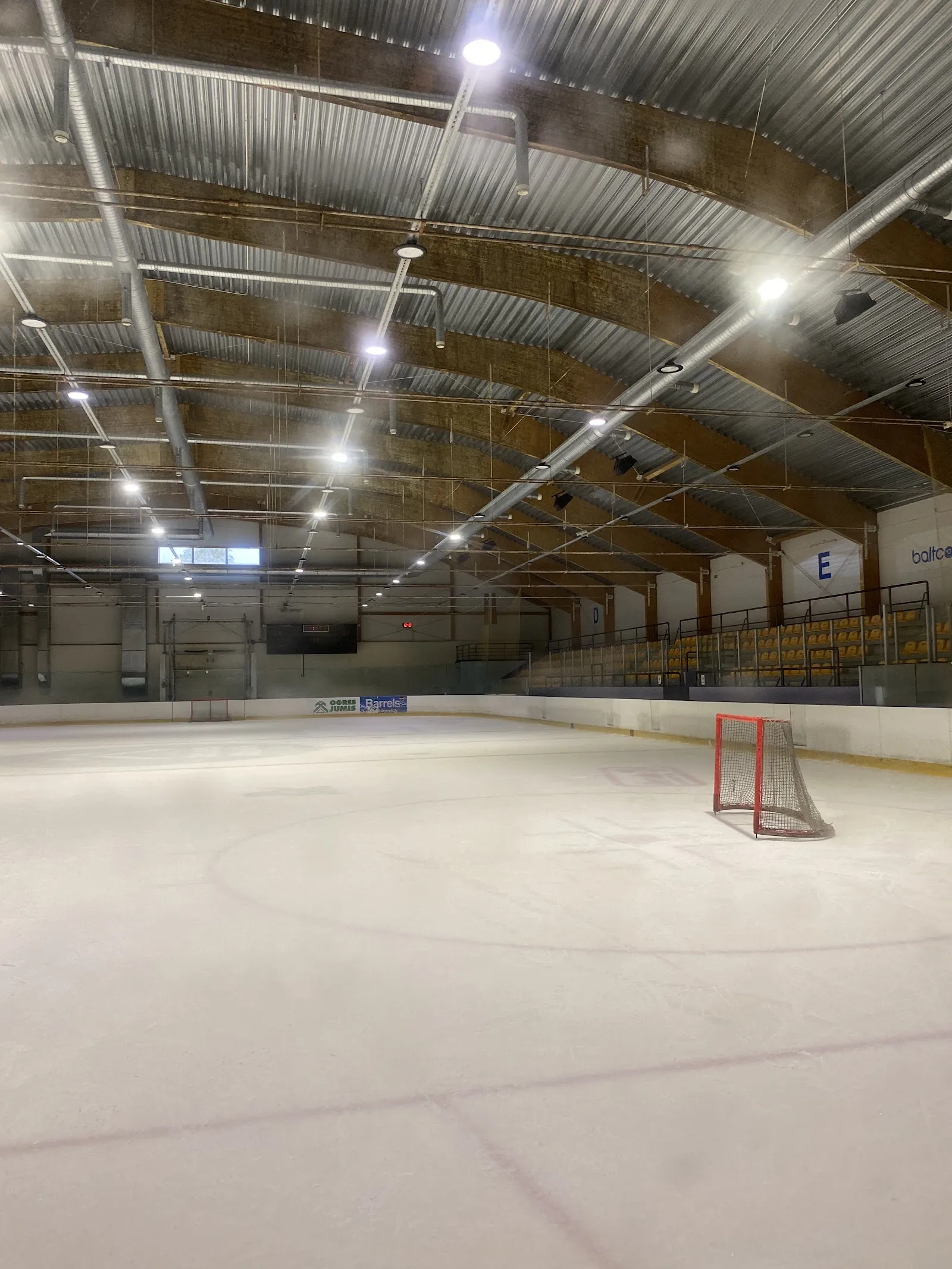 The empty ice sheet under the curved timber roof, with a goal net on the ice and the stands along the far side. — Vidzemes Ledus halle