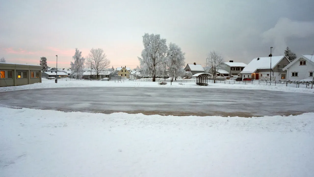 The snow-edged ice surface at Vigernes school at dusk, with a school building on the left and houses beyond. — Vigernes skole skøytebane (Lillestrøm)