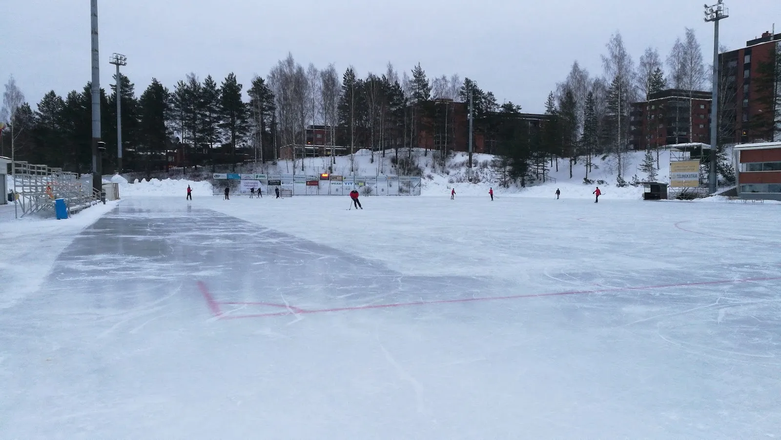 Skaters at a distance on the outdoor artificial ice at Viitaniemi in Jyväskylä, with the marked lines, floodlight masts and pines behind — Viitaniemen ulkotekojäärata