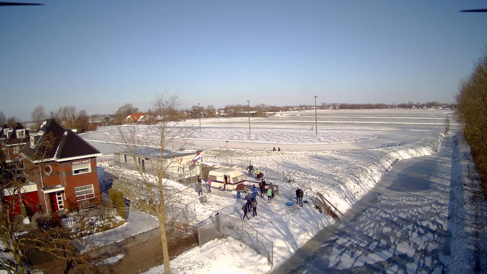 Skaters scattered across the flooded, snow-rimmed Koudekerk natural-ice rink on a clear winter day. — Landijsbaan Koudekerk aan den Rijn (VIJK)