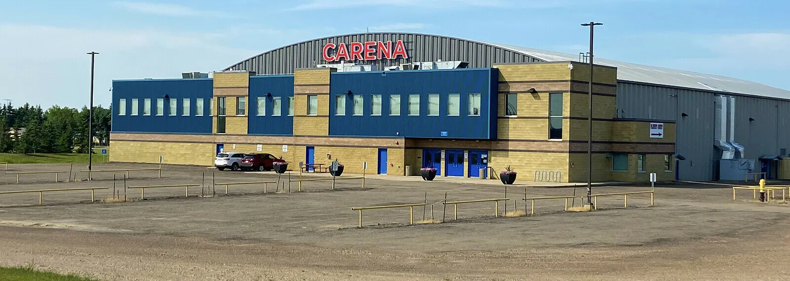 The Viking Carena Complex in summer: a blue and tan two-storey front with the arena's curved roof and the word CARENA above it, behind a gravel parking lot. — Viking Carena Complex