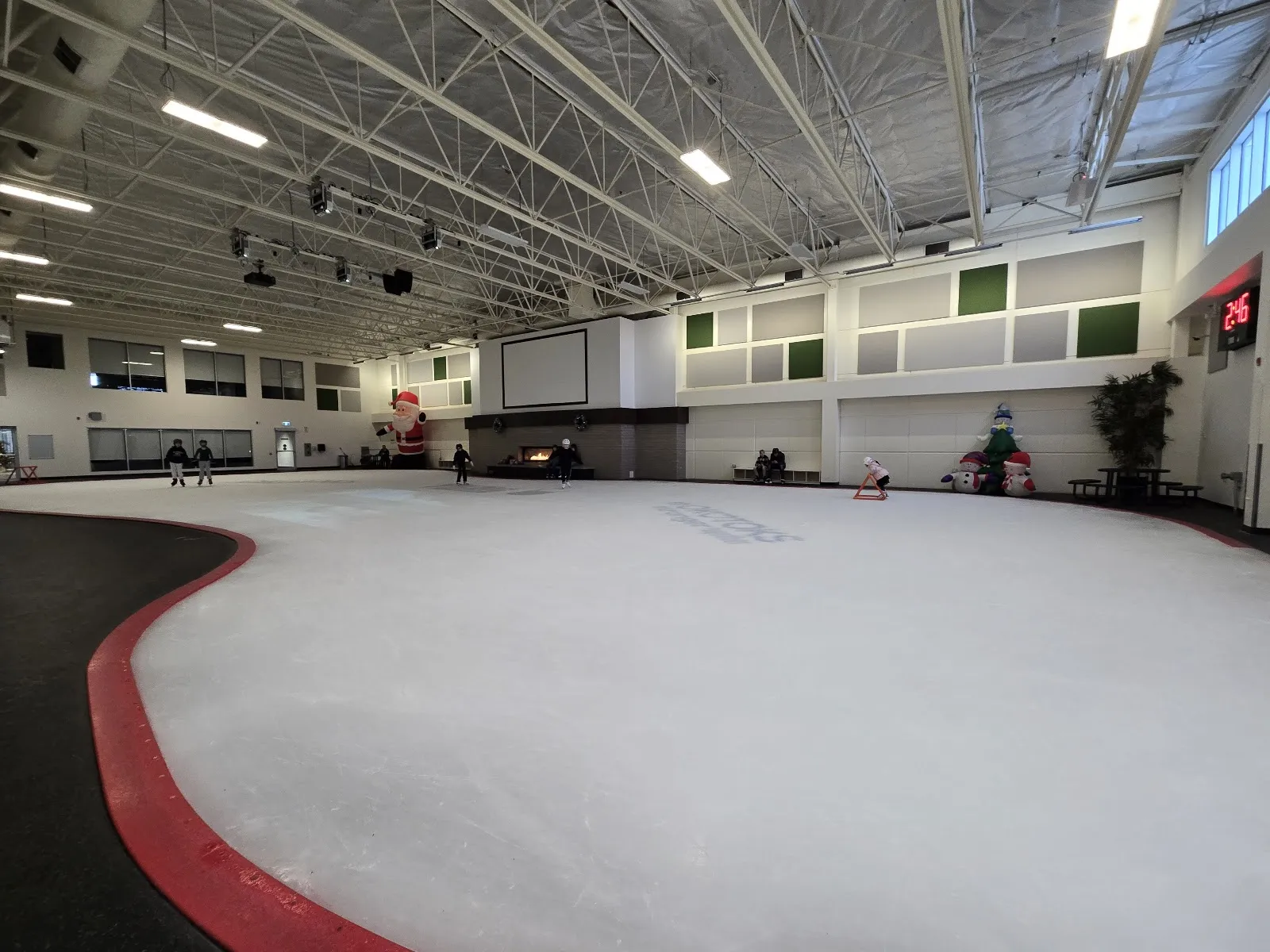 La arena de hielo recreativa: una pista de forma libre con un borde rojo bajo un techo de armaduras blancas, algunos patinadores a lo lejos y adornos festivos a lo largo de la pared del fondo. — Viking Rentals Centre