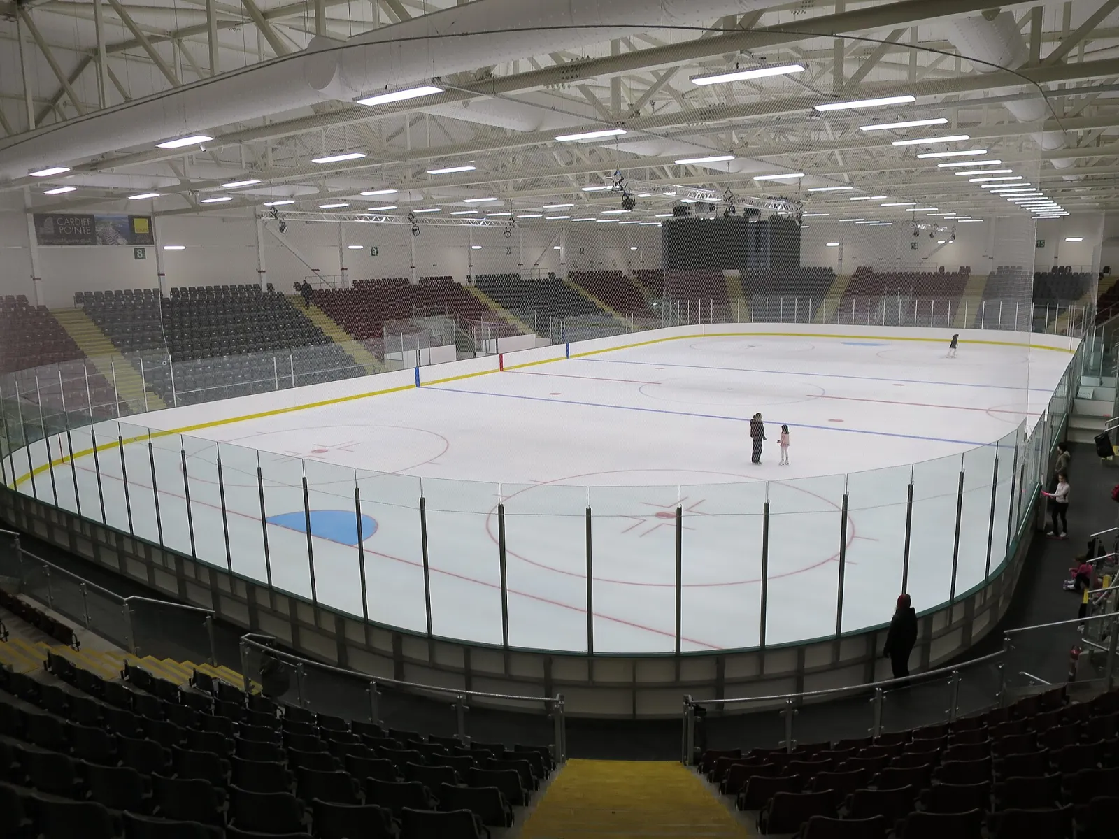 The Olympic-size ice pad from the stands, with the seating bowl and roof trusses above and a few skaters on the ice, February 2016. — Vindico Arena