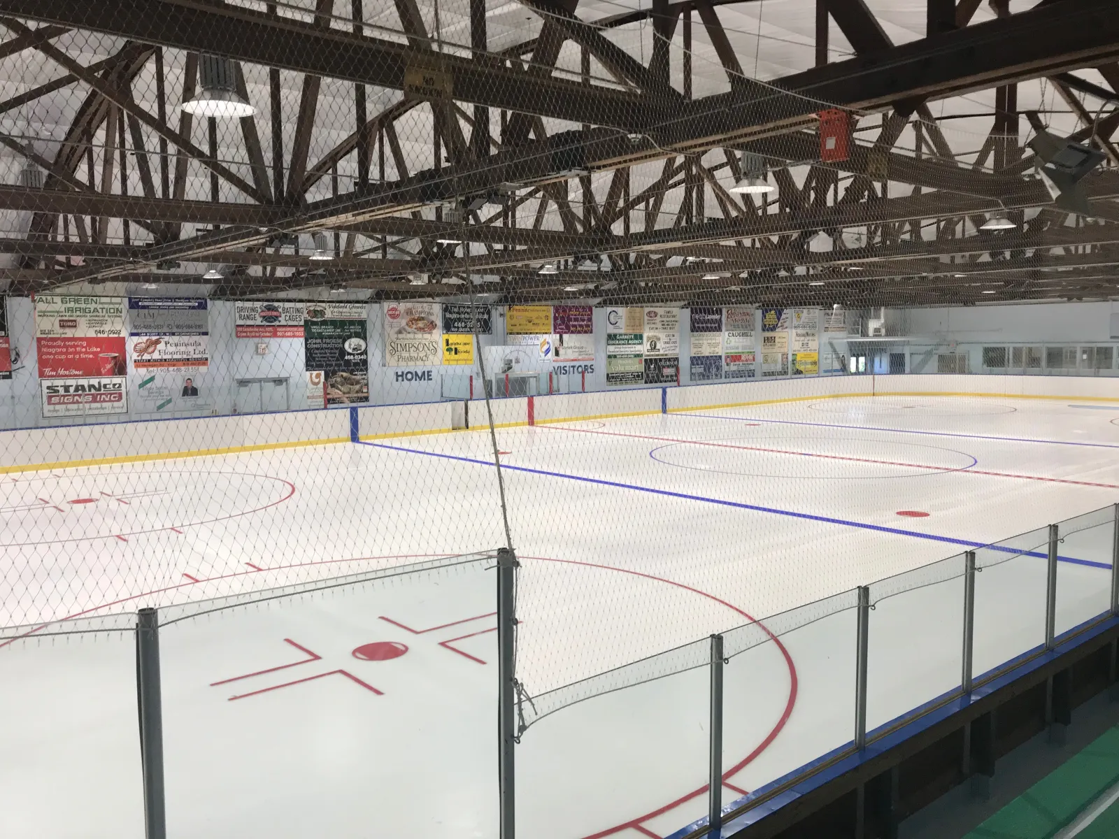 Freshly painted ice in one of the Virgil arenas, seen through the netting behind a goal, with advertising boards along the far wall under a steel truss roof. — Virgil Arenas