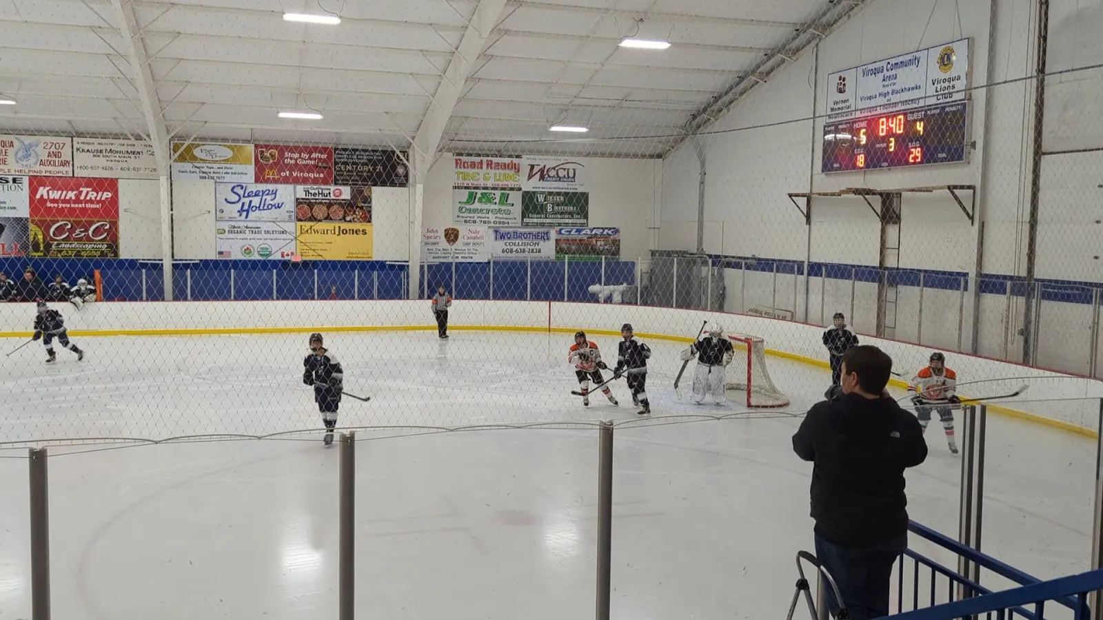 A game on the ice sheet at Viroqua Community Arena, players at the far end beneath the boards advertising — Viroqua Community Arena