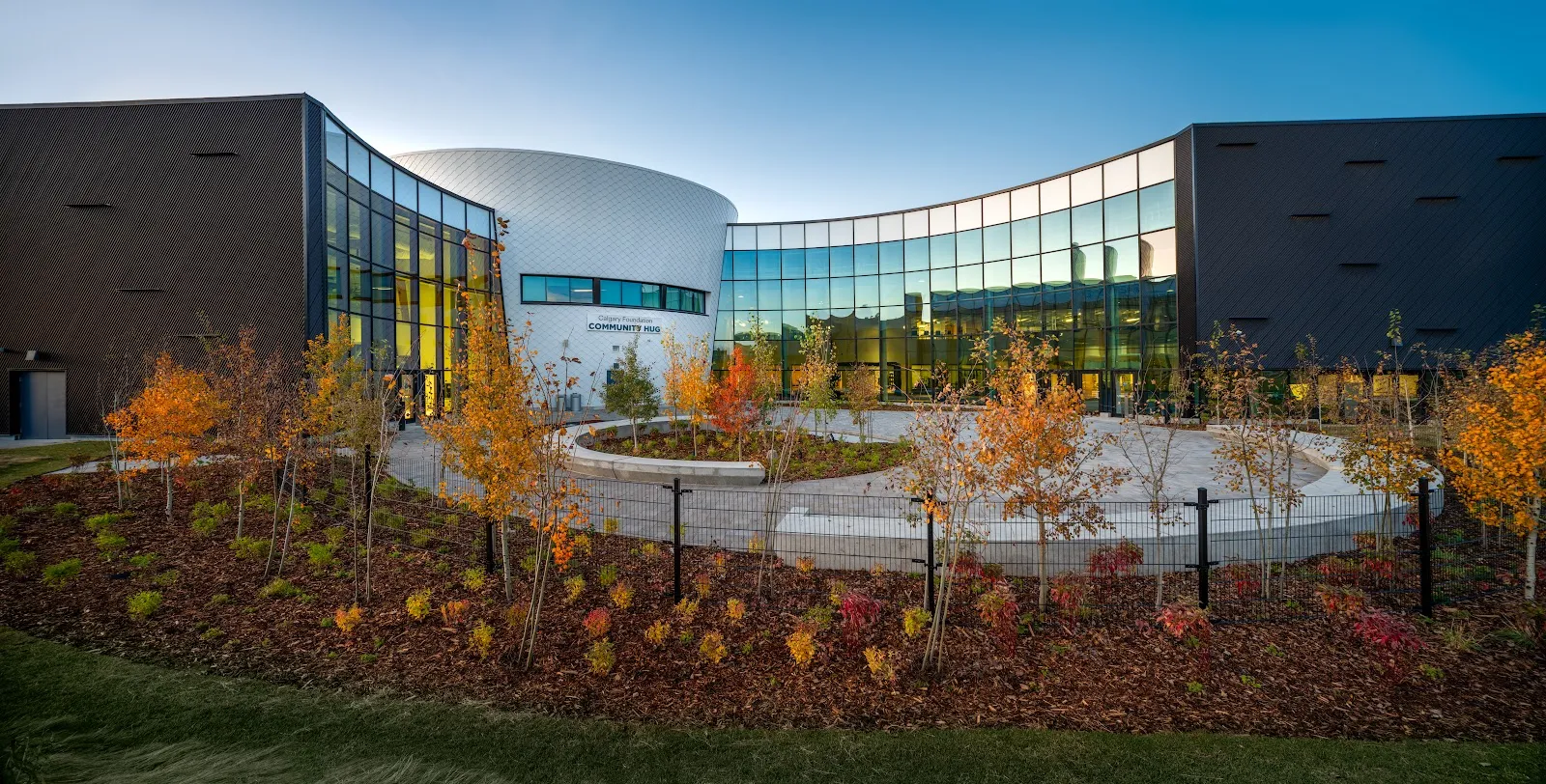 The curved glass entrance between dark-clad wings at dusk, behind a landscaped plaza with autumn trees. — Vivo for Healthier Generations