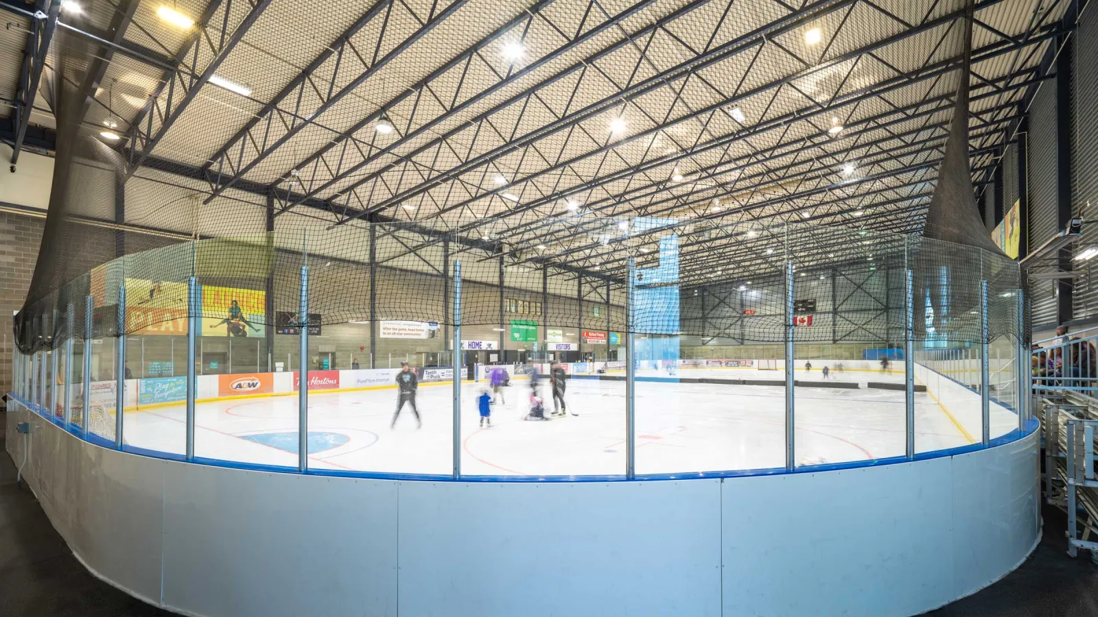 A sheet seen from the corner behind the glass, with a few skaters at centre ice under a steel truss roof and sponsor boards on the far side. — Vivo for Healthier Generations