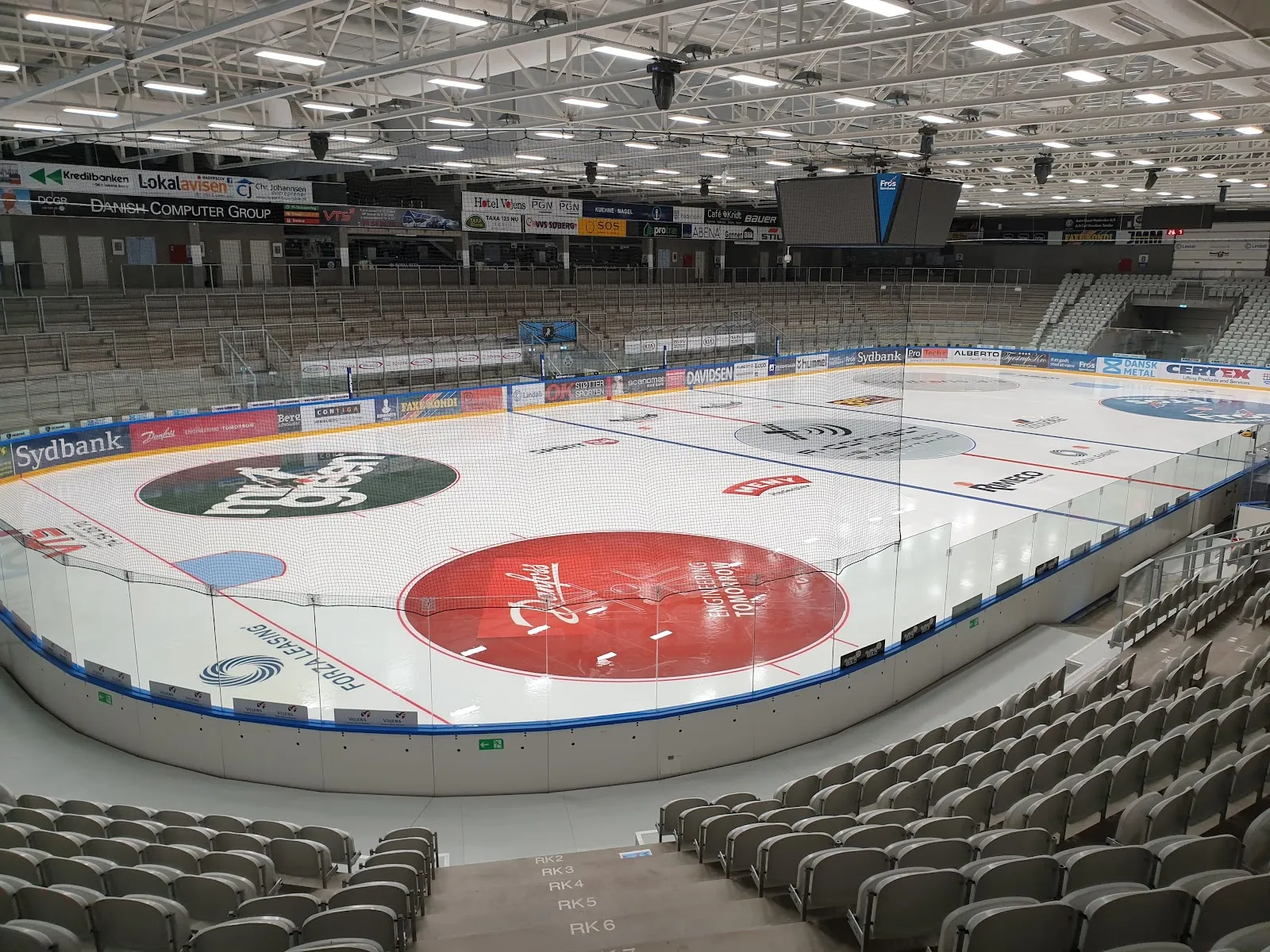 The empty ice sheet of the arena at Stadion Parken in Vojens, seen from the stands with rows of grey seats around the boards and club logos painted on the ice. — Vojens Skøjtehal