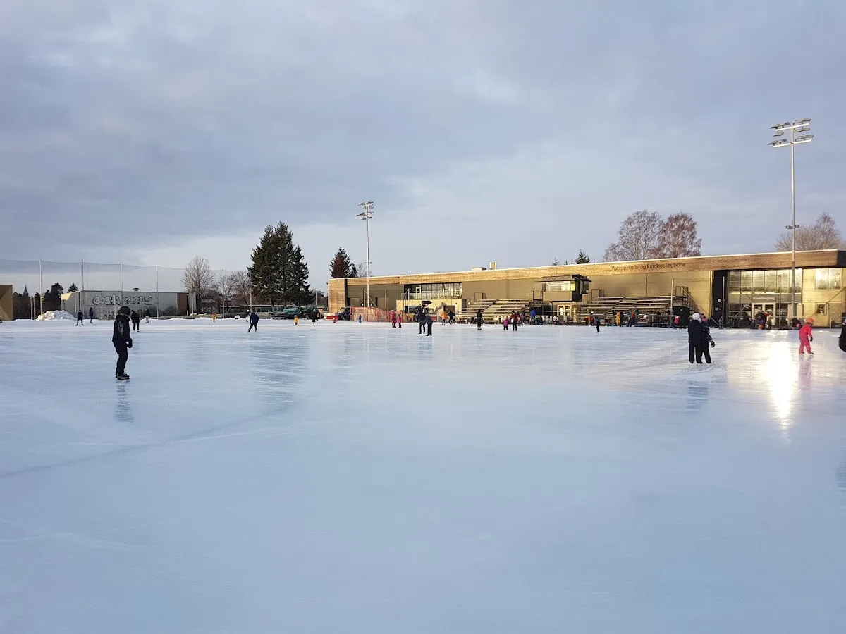 Voldsløkka kunstisbane at dusk: skaters on the ice in front of the clubhouse and floodlights. — Voldsløkka kunstisbane