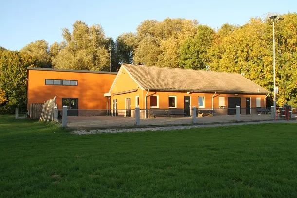 The orange-brick clubhouse of the Volksijsbaan De Eendracht at Genemuiden with a pitched tiled roof, a covered terrace and a floodlight mast, seen across the grass in autumn sun. — Volksijsbaan De Eendracht (Genemuiden)