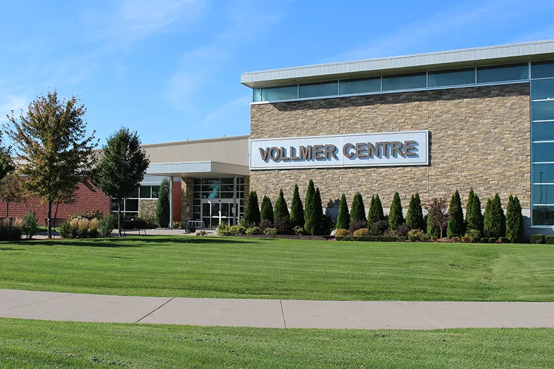 The stone-and-glass front of the building with its 'Vollmer Centre' sign above a row of cedars and a wide lawn. — Vollmer Complex