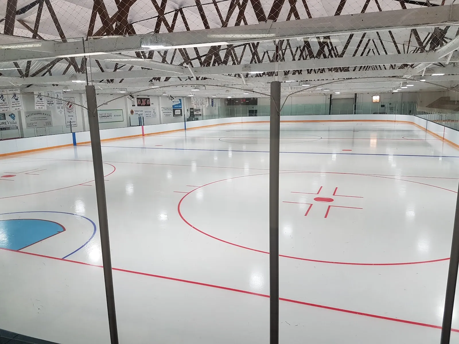 The empty sheet seen through two roof-support posts, with the red centre line, faceoff circles and a blue crease in view under the steel trusses. — Vulcan Arena