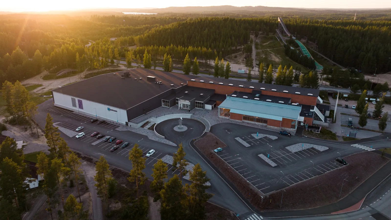 Flygbild över byggnaderna vid Vuokatti Areena med parkering, omgivna av skog och Vuokattis hoppbackar i skymningen – Vuokatti Areena