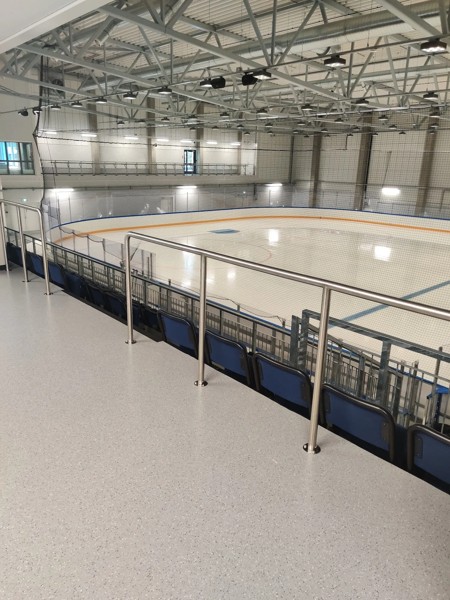 Den tomma isen i Vuokatti Areena sedd från läktaren bakom stålräcket, med sargen och ett tak av stålbalkar ovanför – Vuokatti Areena