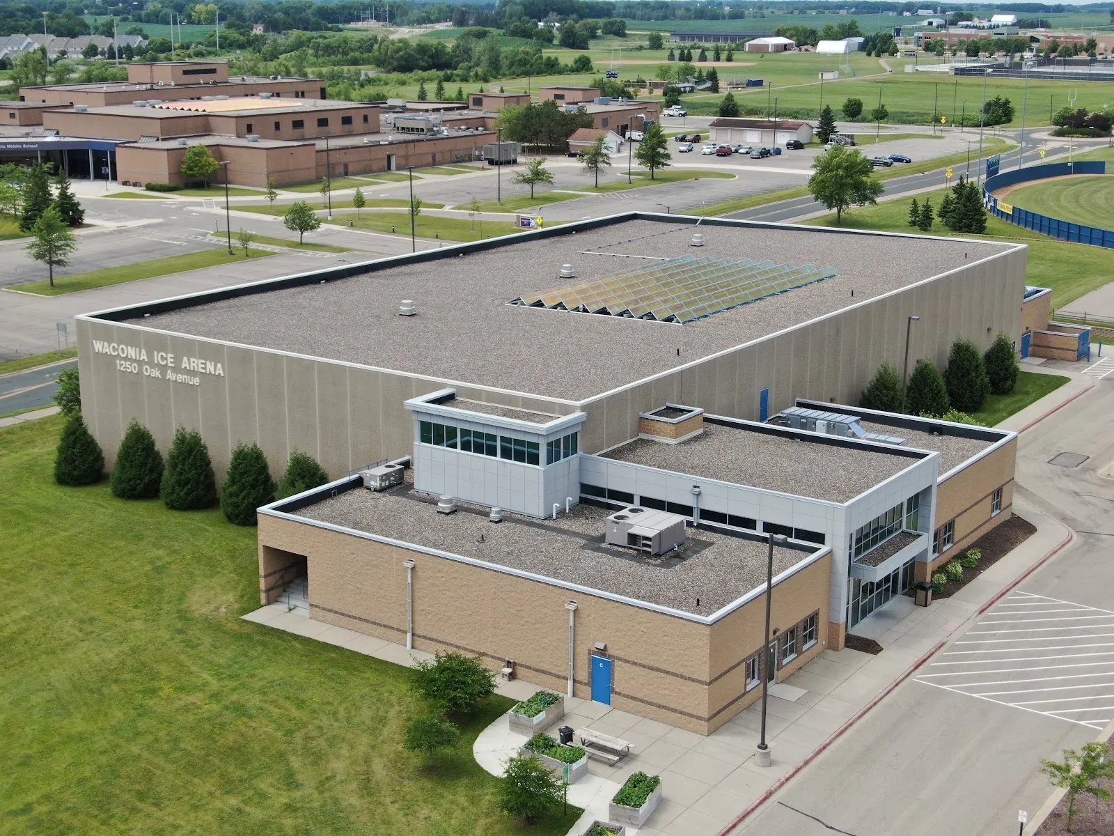 Aerial drone view of the building, with "WACONIA ICE ARENA / 1250 Oak Avenue" painted in large letters on the near wall; parking lot, evergreen trees and the adjoining middle school campus visible around it — Waconia Ice Arena