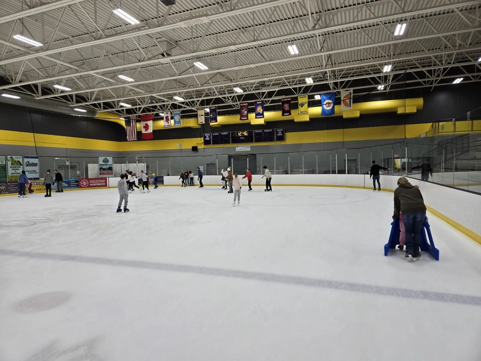 A public session on a wide sheet, skaters spread across the ice and a few skate-aid frames under a steel roof with a black and yellow band and banners on the far wall. — Waconia Ice Arena