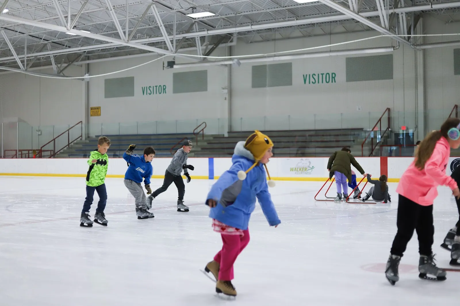 Children skating across a white sheet in front of a grey wall lettered Visitor, a yellow board line along the near side and a steel-framed roof overhead. — Walker Ice and Fitness Center