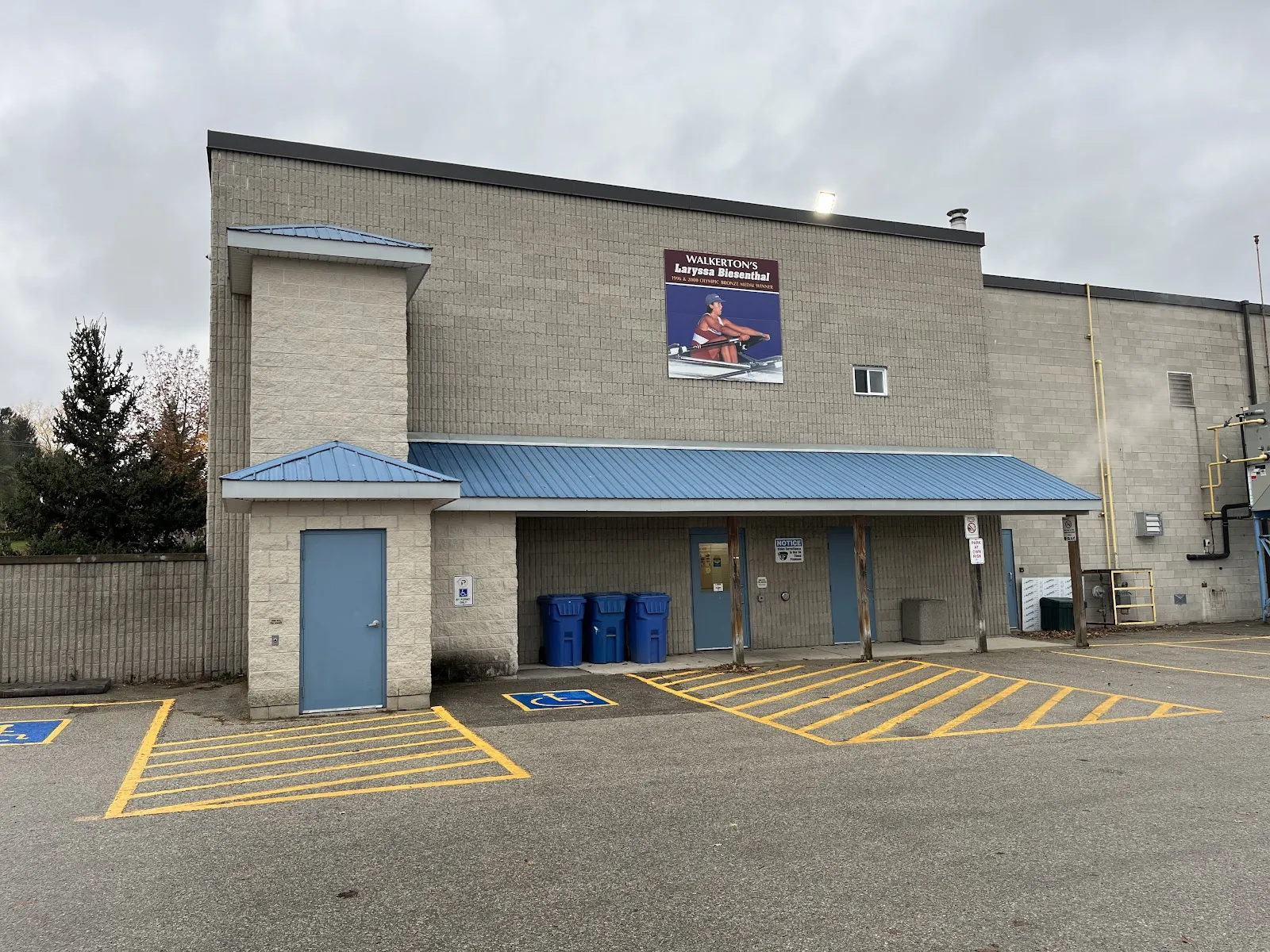 The tan brick side of the Walkerton Community Centre with its blue canopy, side doors and accessible parking spaces, seen from the parking lot. — Walkerton Community Centre
