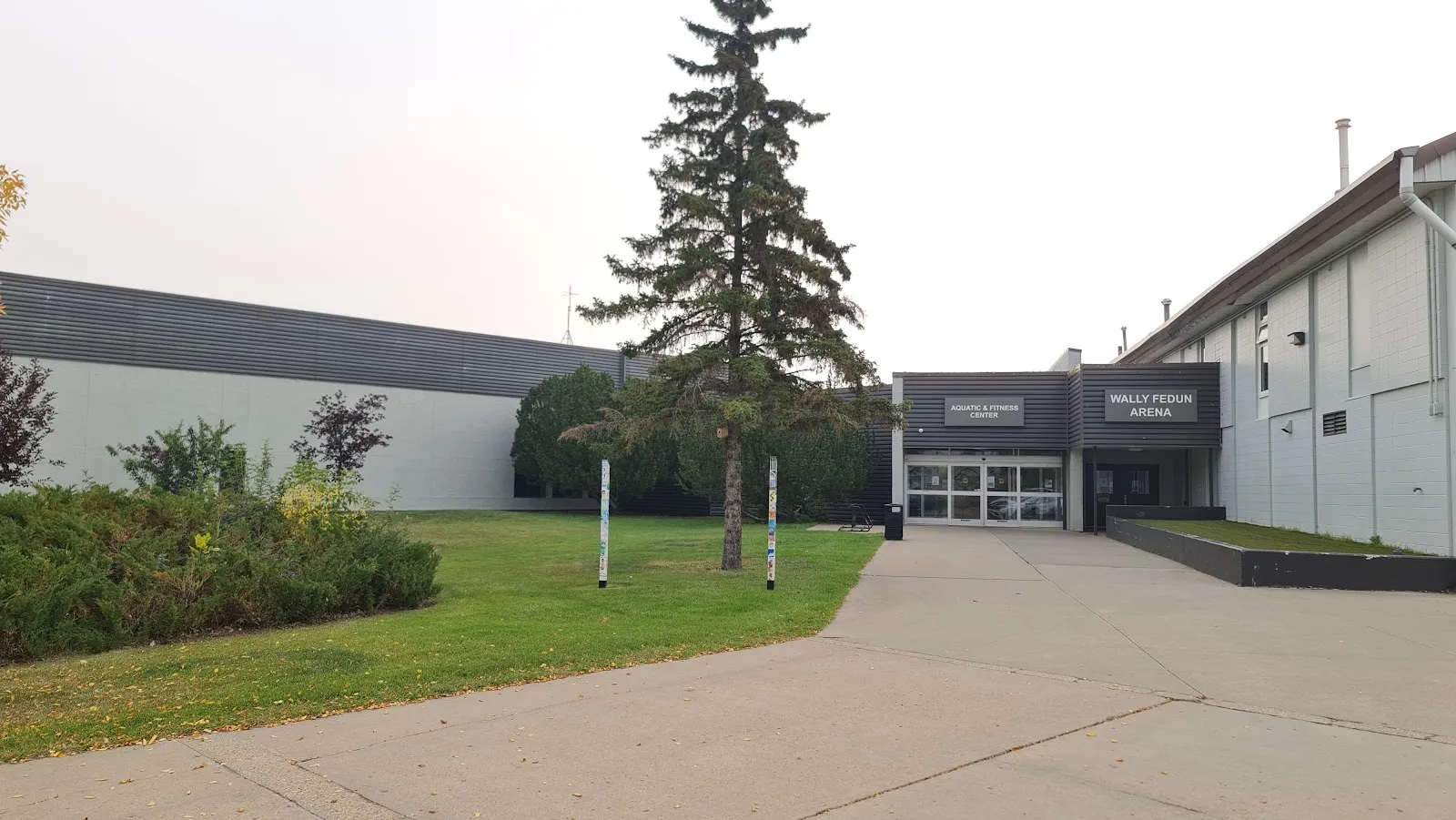 La baja entrada gris del Vegreville Recreation Centre, con letreros de Aquatic & Fitness Center y Wally Fedun Arena, detrás de un abeto y pasto. — Wally Fedun Arena