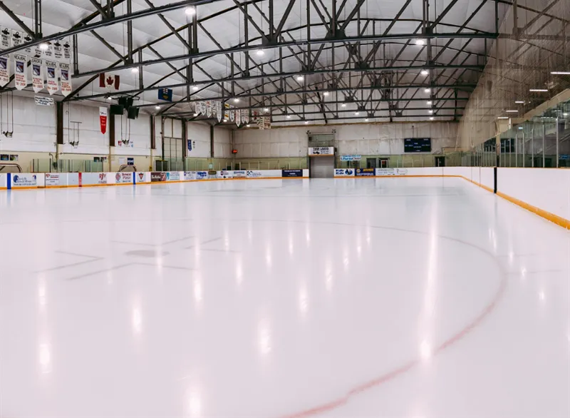 El hielo vacío de Wally Fedun Arena bajo armaduras de acero en el techo, con mantas de equipos en el muro cercano y el marcador en el extremo opuesto. — Wally Fedun Arena