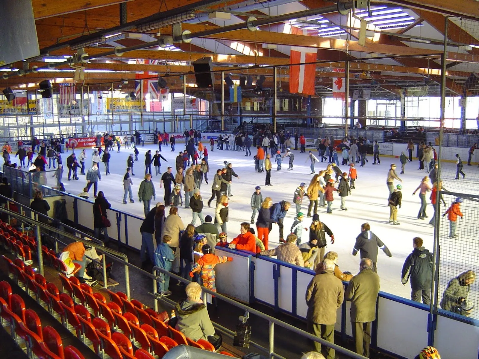 A crowded public session at the Walter-Maack ice stadium, skaters of all ages across the ice with red stand seating along the boards and the timber roof overhead. — Walter-Maack-Eisstadion