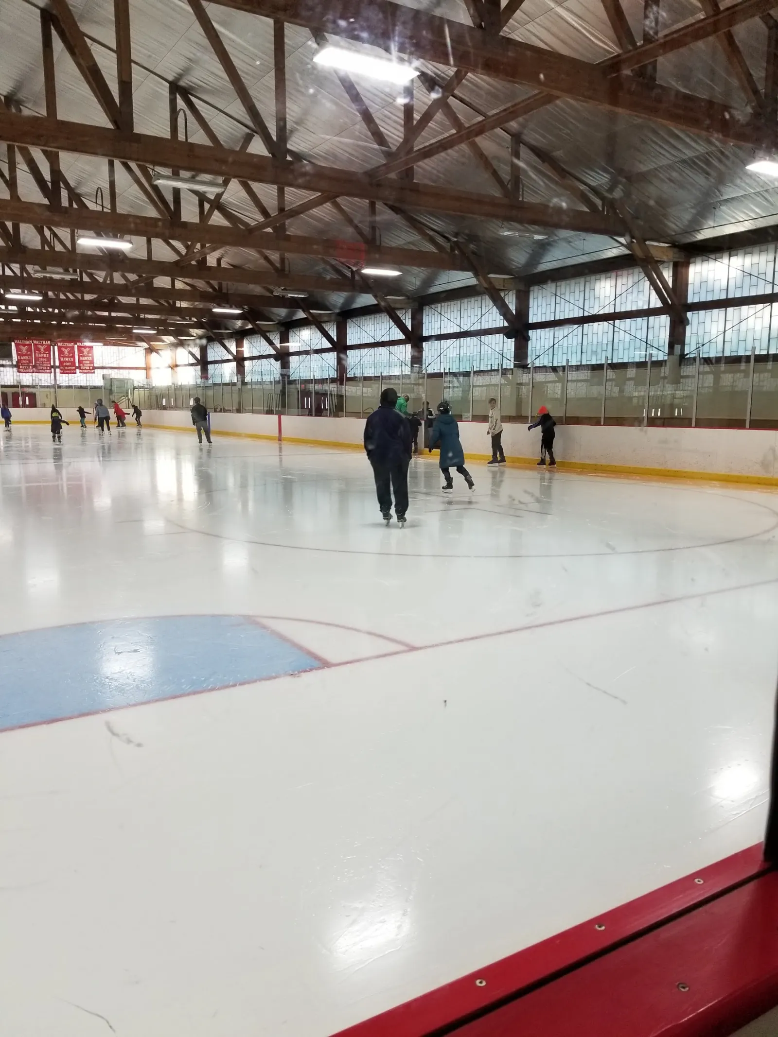 Patinadores del público repartidos por el hielo bajo las oscuras armaduras de madera del techo de la pista, con una hilera de ventanas altas a lo largo del lado opuesto. — Waltham Veterans Memorial Rink