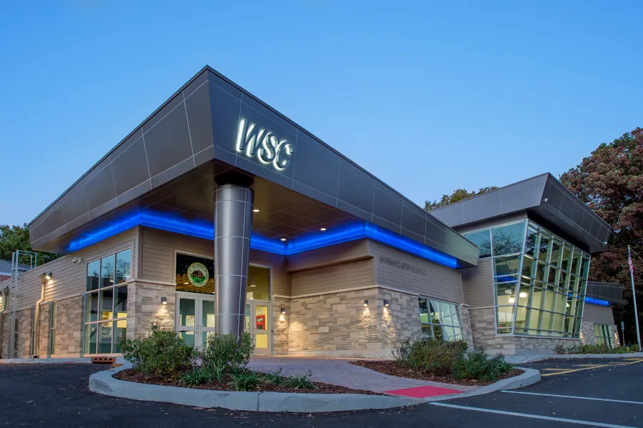 La entrada renovada del Warinanco Sports Center al anochecer, con un letrero iluminado WSC en la marquesina inclinada, muros de piedra y vidrio e iluminación de acento azul. — Warinanco Sports Center