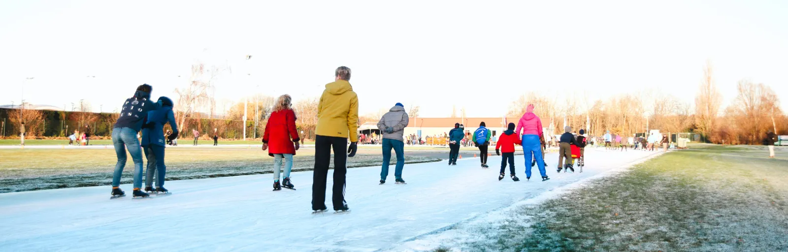 Een groep schaatsers op een strook natuurijs op de landijsbaan van Warmond in december 2022 in lage winterzon, met grasranden aan beide kanten en bomen langs de overkant. – Landijsbaan Warmond (Warmondse IJs- en Skeelerclub)
