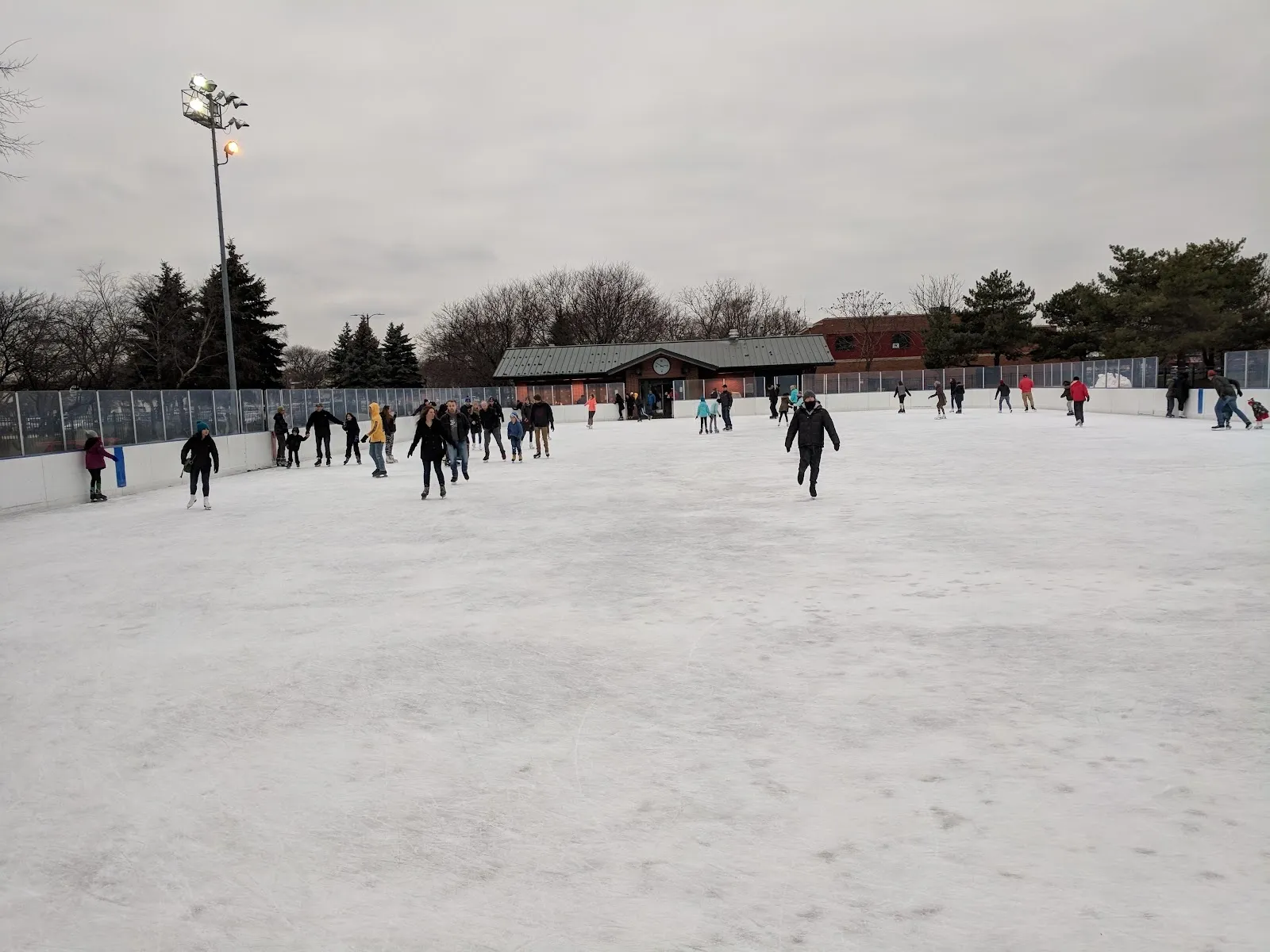 The Warren Park rink under a grey sky, skaters spread across a wide flooded oval with a lodge roof and trees at the far edge. — Warren Park Ice Rink