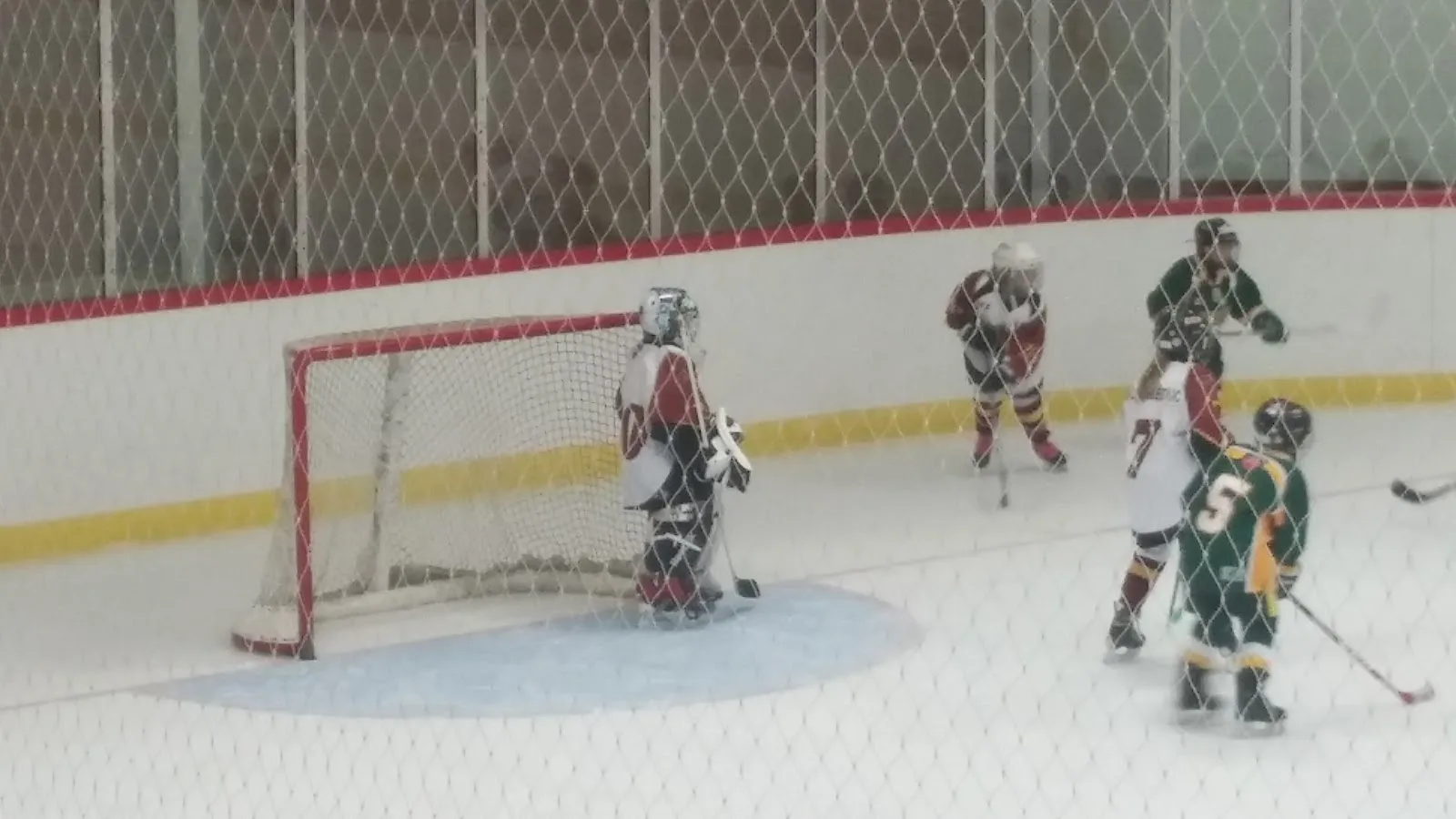 Youth players around the net in a game at the Warsaw Community Centre arena, seen through the rink fence. — Warsaw Community Centre