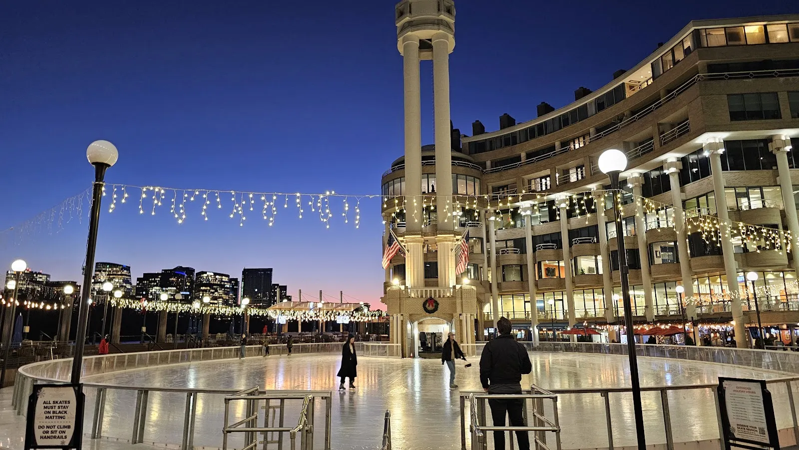 The circular Washington Harbour rink at dusk with a few skaters, the tower and the waterfront buildings behind and string lights overhead — Washington Harbour Ice Rink