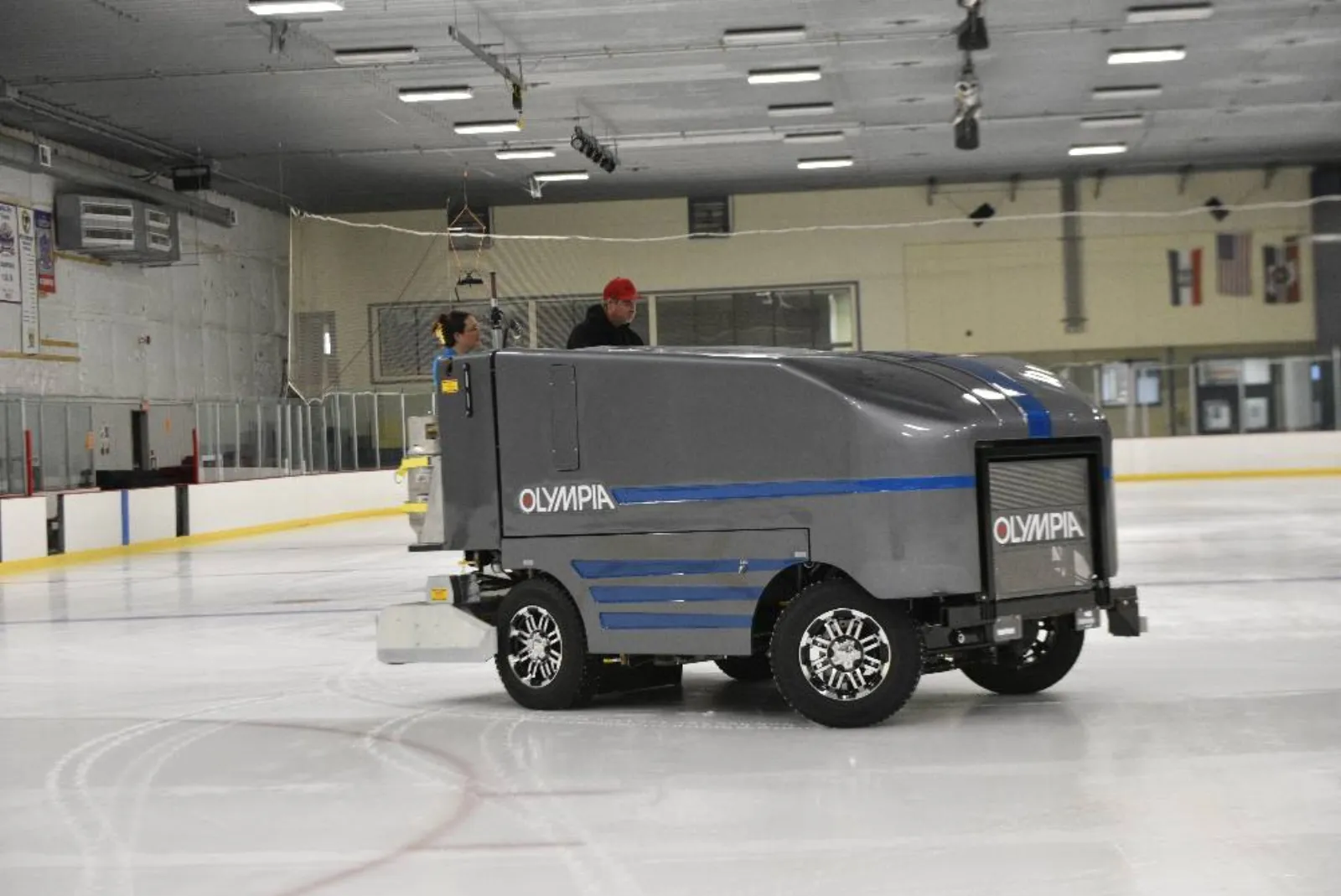 Una pulidora de hielo Olympia recorre la pista, con las vallas, banderas en la pared y la iluminación del techo visibles. — Washington Park Ice Arena