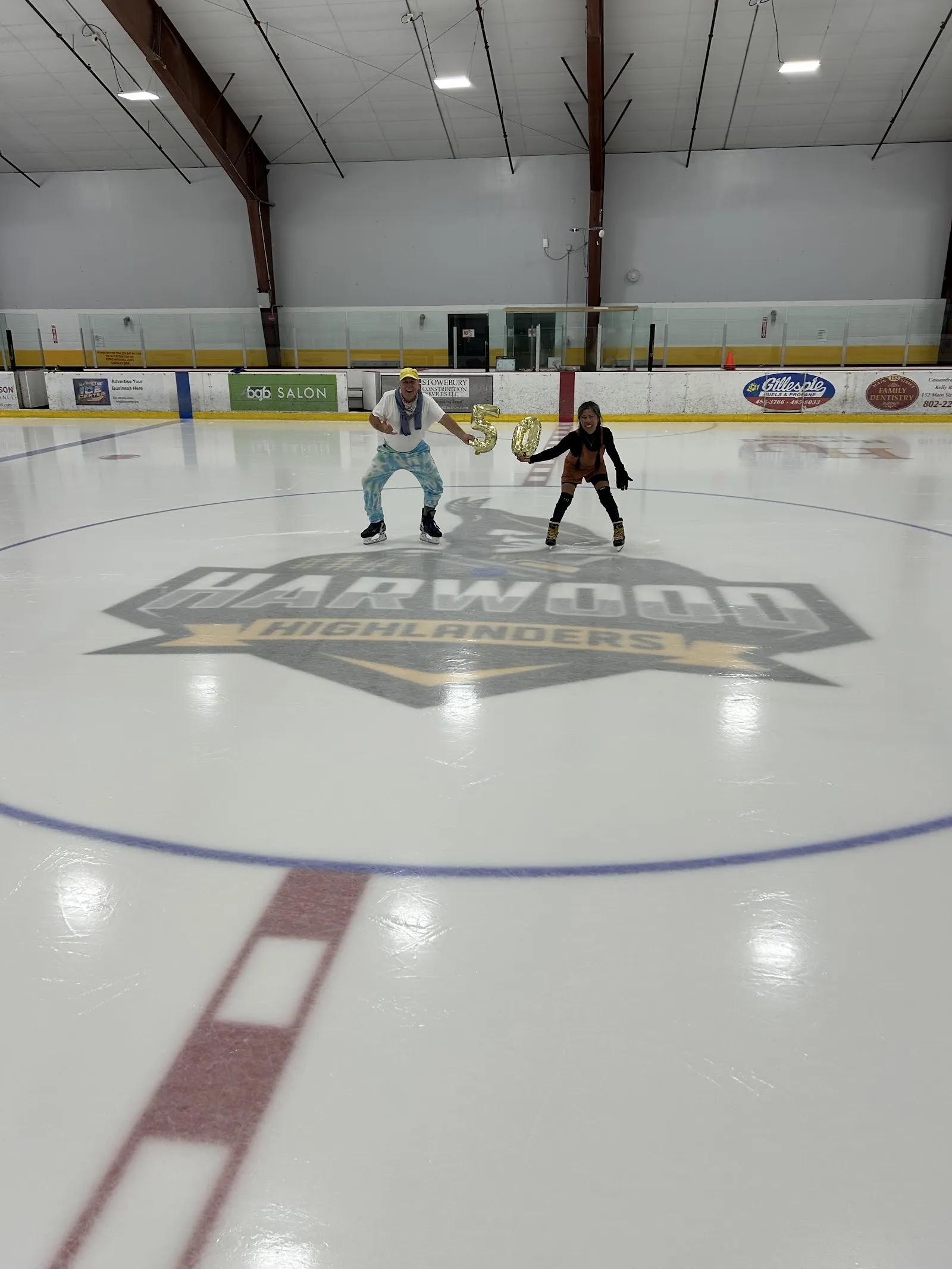 La pista con un tenue logotipo en el centro y dos personas cerca del centro del hielo, un techo con estructura de acero y anuncios de patrocinadores en la pared del fondo. — Waterbury Ice Center
