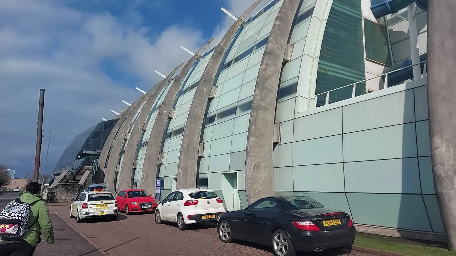 The curved glass-and-concrete side of the Waterfront leisure building on the Greenock seafront, with cars parked along the pavement. — Waterfront Ice Rink