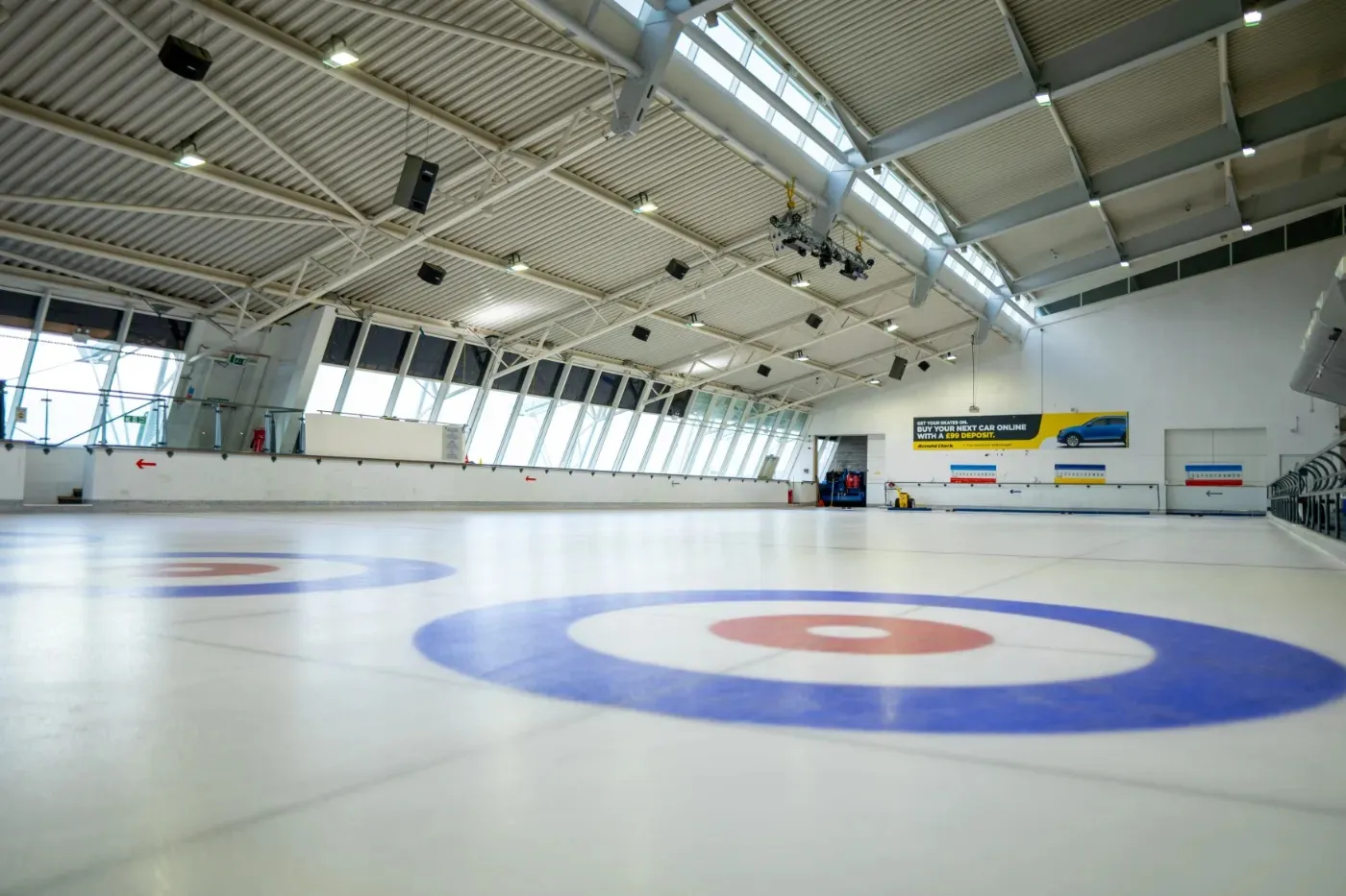 The empty Waterfront rink hall at Greenock, with a curling target painted on the ice and the long glazed side wall and steel roof beyond. — Waterfront Ice Rink
