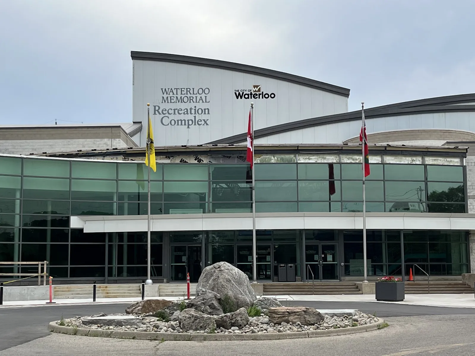La entrada con frente de vidrio del Waterloo Memorial Recreation Complex, con su nombre en el muro superior curvo, astas de bandera y un jardín de rocas al frente — Waterloo Memorial Recreation Complex