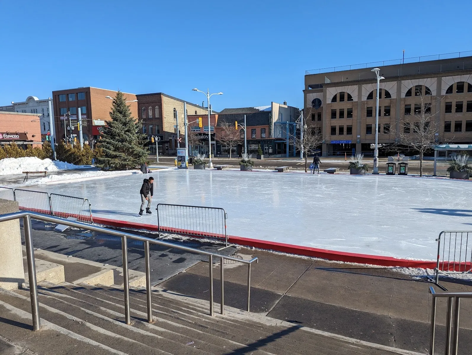 The Waterloo Public Square rink in summer light, with a skater on the ice and the uptown buildings behind — Waterloo Public Square Rink