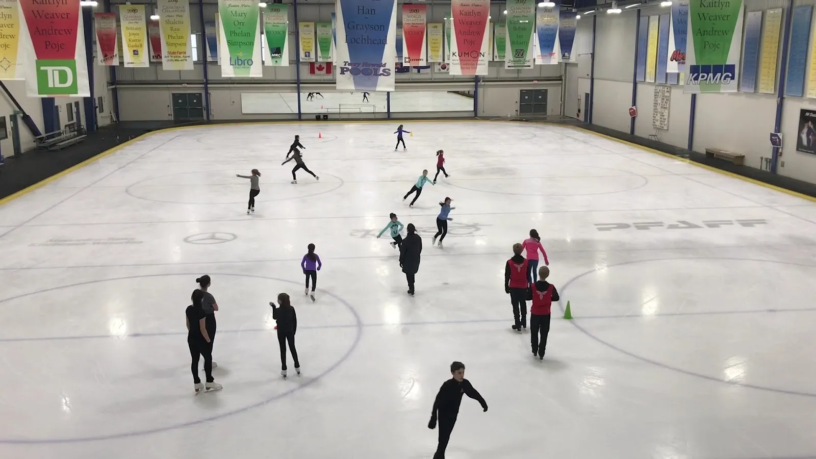 A public skating session under a roof with coloured pennants, skaters spread across a wide sheet seen from the glass. — RIM Park
