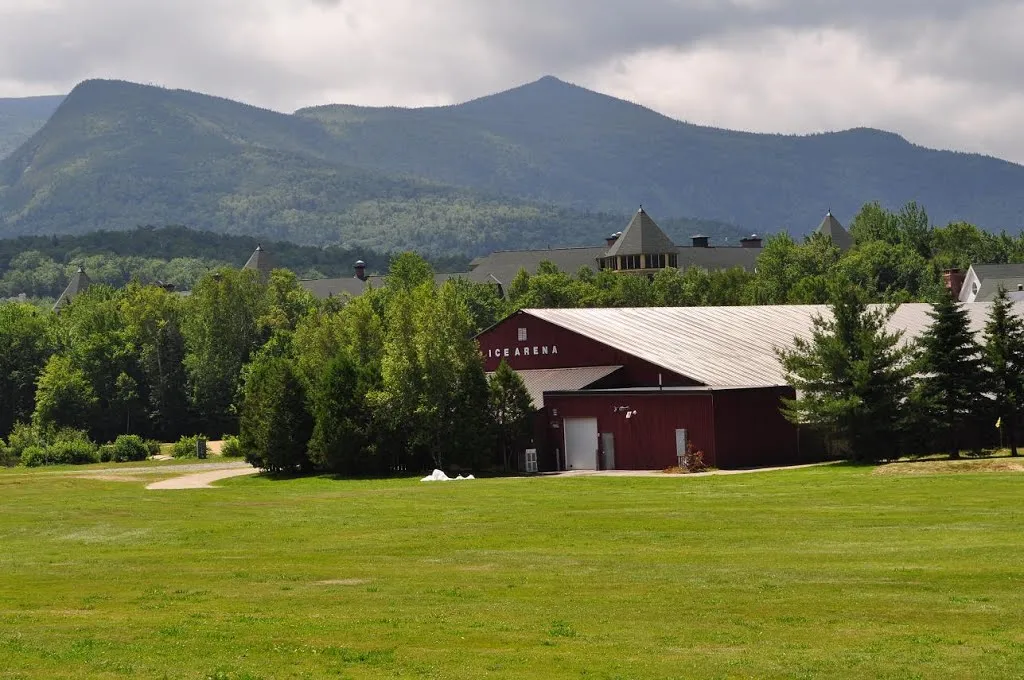 El edificio de la arena con revestimiento metálico rojo oscuro rotulado ICE ARENA, visto desde el otro lado de un pasto abierto con las White Mountains detrás. — Waterville Valley Ice Arena