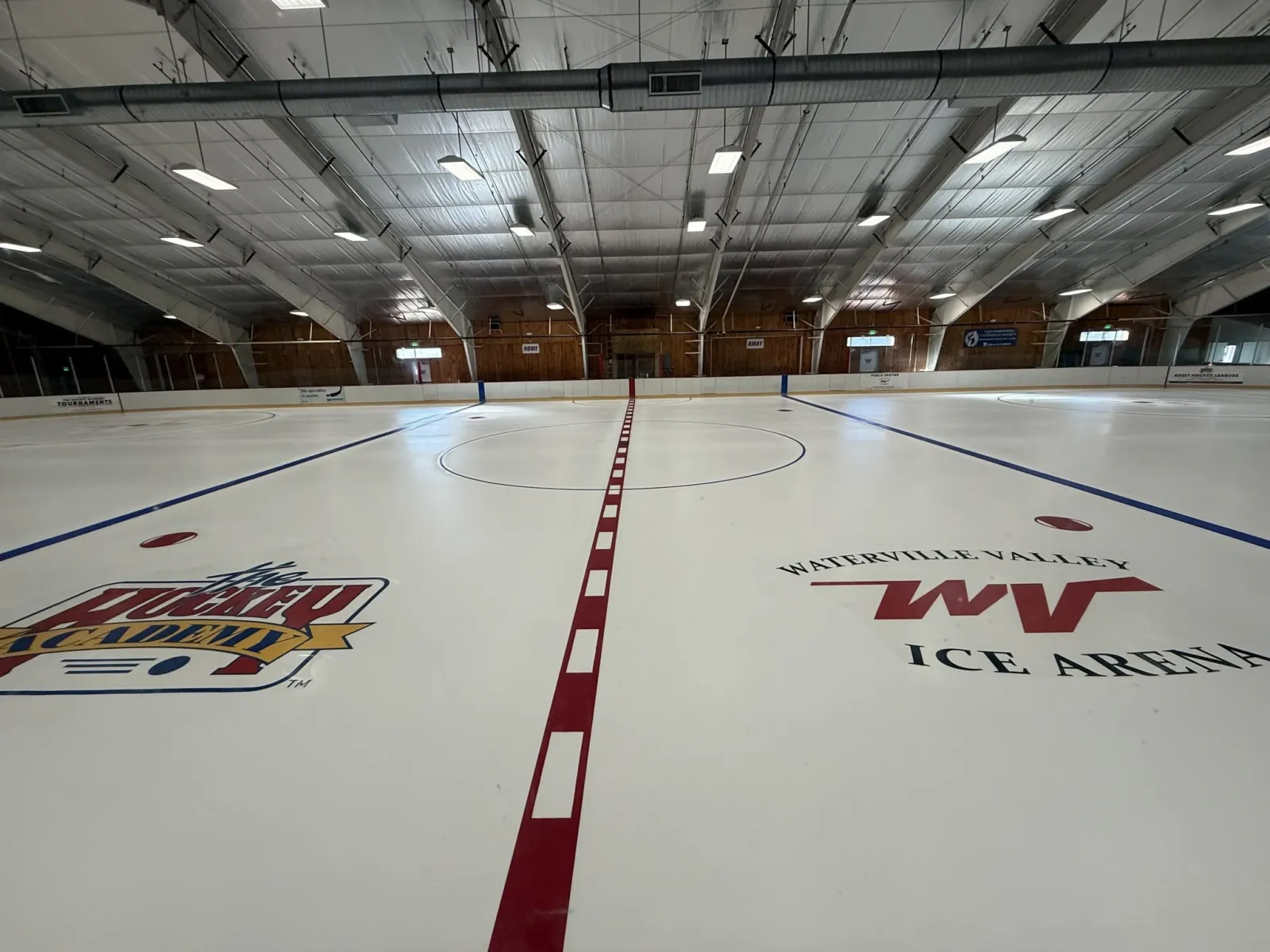 Pista vacía recién pulida desde el nivel del hielo, con el logotipo "WV" de Waterville Valley Ice Arena y el logotipo de The Hockey Academy pintados en el centro de la pista, vallas del fondo con paneles de madera y armaduras de acero en el techo. — Waterville Valley Ice Arena