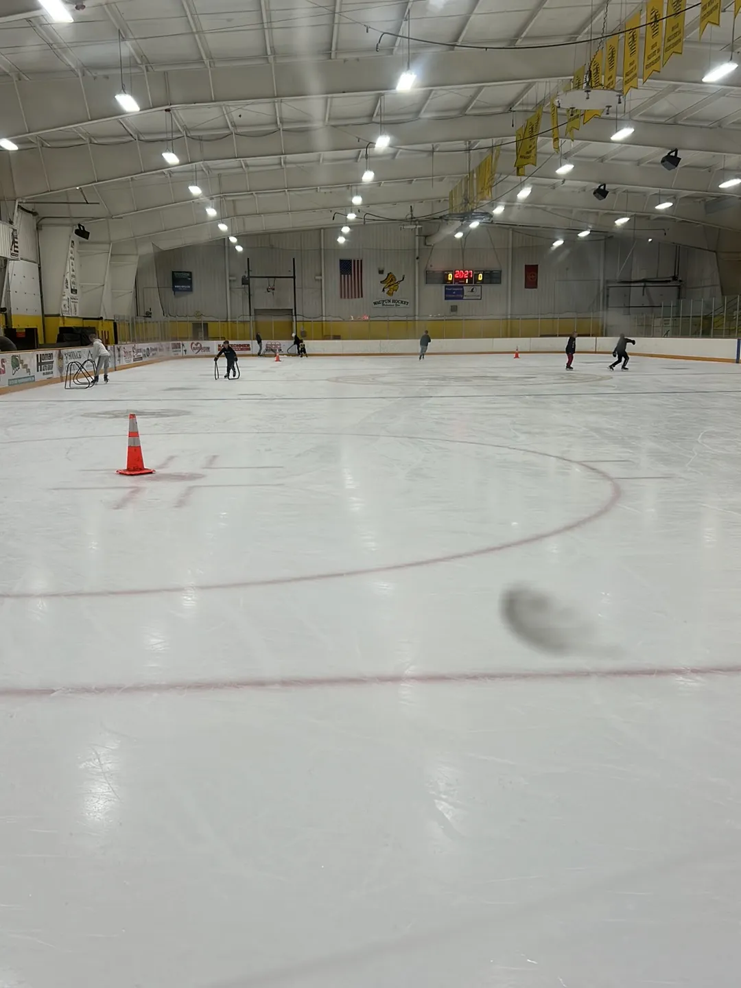 The ice seen from centre, with a cone and a few skaters, yellow championship banners along the roof and flags at the far end. — Waupun Ice Arena