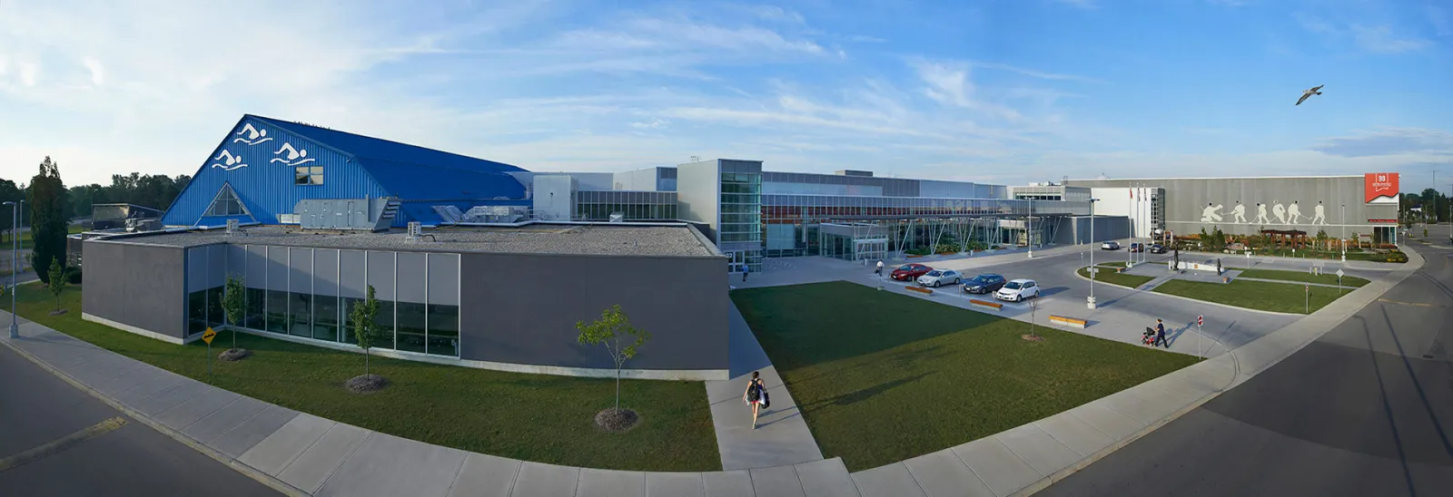 Wide view of the Wayne Gretzky Sports Centre on a clear evening: the blue peaked roof over the pool wing, the glass entrance block and the long grey arena walls, with lawns and a parking lot in front. — Wayne Gretzky Sports Centre