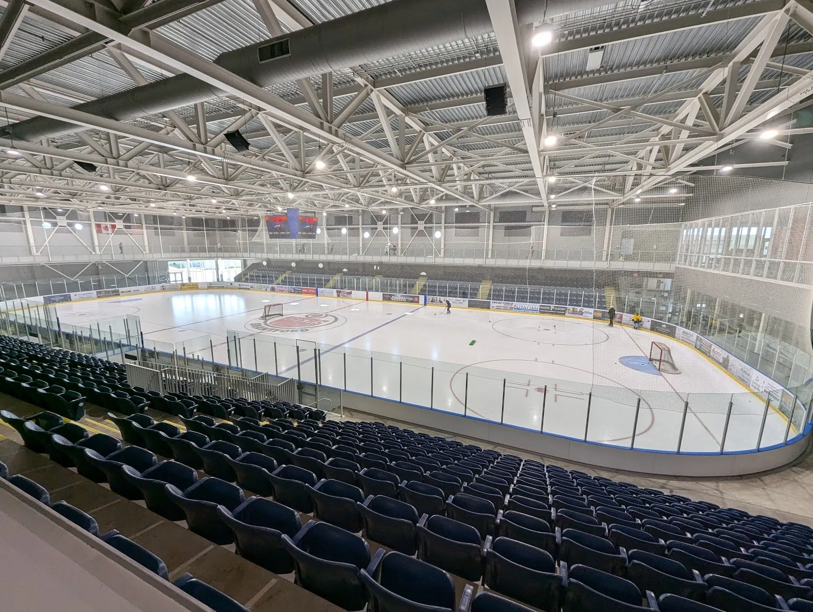 A wide empty sheet behind rows of dark blue seats, white boards and a steel-trussed ceiling above. — Wayne Gretzky Sports Centre