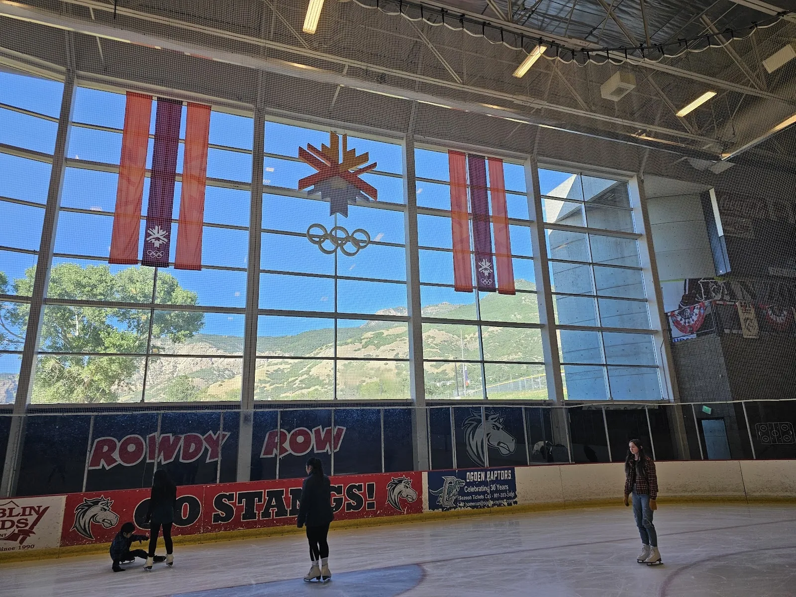 La pista de aspecto vacío vista desde las gradas bajo un alto techo de madera y acero, con una alta pared de ventanas de la que cuelgan banderines olímpicos en el lado opuesto. — Weber County Ice Sheet